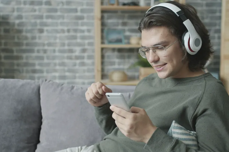 Ein junger Mann mit Kopfhörern sitzt auf dem Sofa und lächelt auf sein Handy.